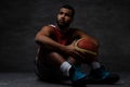 Young African-American basketball player in sportswear sitting on a floor with a ball on a dark background. Royalty Free Stock Photo