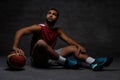 Young African-American basketball player in sportswear sitting on a floor with a ball on a dark background. Royalty Free Stock Photo