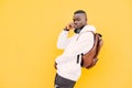 Young african American black man against a yellow wall wearing a white sweatshirt and a backpack listening music on headphones Royalty Free Stock Photo
