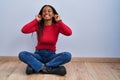 Young african american with braids sitting on the floor at home smiling pulling ears with fingers, funny gesture Royalty Free Stock Photo