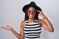 Young african american with braids wearing hat and sunglasses confused and annoyed with open palm showing copy space and pointing