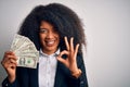 Young african american business woman with afro hair holding cash dollars banknotes doing ok sign with fingers, excellent symbol Royalty Free Stock Photo