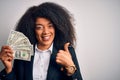 Young african american business woman with afro hair holding cash dollars banknotes happy with big smile doing ok sign, thumb up Royalty Free Stock Photo