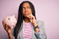 Young african american business woman saving money on piggy bank over background serious face thinking about question,