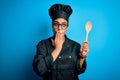 Young african american chef girl wearing cooker uniform and hat holding wooden spoon tool cover mouth with hand shocked with shame