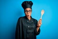 Young african american chef girl wearing cooker uniform and hat holding wooden spoon tool scared in shock with a surprise face,