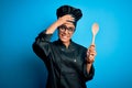 Young african american chef girl wearing cooker uniform and hat holding wooden spoon tool stressed with hand on head, shocked with