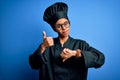 Young african american chef woman wearing cooker uniform and hat over blue background Doing thumbs up and down, disagreement and Royalty Free Stock Photo