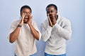 Young african american couple standing over blue background together shouting angry out loud with hands over mouth Royalty Free Stock Photo
