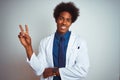 Young african american doctor man wearing coat standing over isolated white background smiling with happy face winking at the Royalty Free Stock Photo