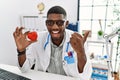 Young african american doctor man wearing doctor uniform holding heart at the clinic pointing thumb up to the side smiling happy Royalty Free Stock Photo