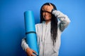 Young african american fitness woman holding yoga mat over isolated blue background stressed with hand on head, shocked with shame Royalty Free Stock Photo