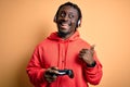 Young african american gamer man playing video game using joystick and headphones pointing and showing with thumb up to the side Royalty Free Stock Photo