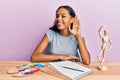 Young african american girl artist sitting at studio table smiling with hand over ear listening and hearing to rumor or gossip Royalty Free Stock Photo