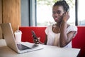 Young African American girl sitting in restaurant with laptop. Pretty girl with earphones joyfully looking in laptop at cafe. Royalty Free Stock Photo