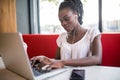 Young African American girl sitting in restaurant with laptop. Pretty girl with earphones joyfully looking in laptop at cafe. Royalty Free Stock Photo