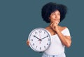 Young african american girl holding big clock serious face thinking about question with hand on chin, thoughtful about confusing Royalty Free Stock Photo