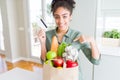 Young african american girl holding paper bag of groceries and credit card as payment with surprise face pointing finger to Royalty Free Stock Photo