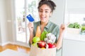 Young african american girl holding paper bag of groceries and credit card as payment very happy pointing with hand and finger to Royalty Free Stock Photo