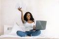 Young african american girl woman in white t-shirts sitting on bed with laptop in bedroom, holding notebook showing to Royalty Free Stock Photo