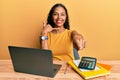 Young african american girl working at the office with laptop and calculator smiling doing talking on the telephone gesture and Royalty Free Stock Photo