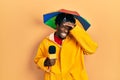 Young african american journalist man wearing yellow raincoat and umbrella cap stressed and frustrated with hand on head,