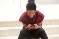 Young african american man with beanie sitting on steps outside and looking at mobile phone Royalty Free Stock Photo