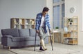 Young African American man with a broken leg in a cast walking with crutches at home