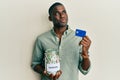 Young african american man holding credit card and jar with dollars smiling looking to the side and staring away thinking Royalty Free Stock Photo