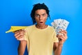 Young african american man holding paper plane and dollars in shock face, looking skeptical and sarcastic, surprised with open Royalty Free Stock Photo