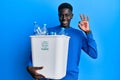 Young african american man holding recycling wastebasket with plastic bottles doing ok sign with fingers, smiling friendly Royalty Free Stock Photo