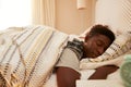 Young African American man lying asleep in bed in the morning, alarm clock in the foreground, selective focus