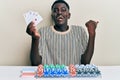 Young african american man playing poker holding cards pointing thumb up to the side smiling happy with open mouth