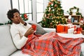 Young african american man sitting on the sofa drinking coffee by christmas tree smelling something stinky and disgusting,