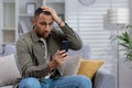 Young african american man sitting on sofa at home and looking at phone in shock, holding hand on head Royalty Free Stock Photo