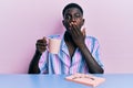 Young african american man sitting on the table reading a book and drinking a coffee covering mouth with hand, shocked and afraid Royalty Free Stock Photo