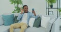 Young African American man with smartphone making video call while sitting on couch in living room. Portrait of happy Royalty Free Stock Photo