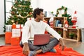 Young african american man using laptop sitting by christmas tree looking away to side with smile on face, natural expression Royalty Free Stock Photo