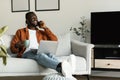 Young african american man using laptop and talking on mobile phone, drinking coffee, working remotely from home Royalty Free Stock Photo