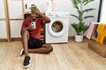 Young african american man using smartphone waiting for washing machine surprised with hand on head for mistake, remember error