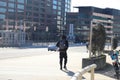 A young African American man wearing a black hoodie walking along a wide smooth sidewalk along a street with glass office building