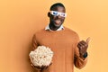 Young african american man wearing d glasses holding popcorns pointing thumb up to the side smiling happy with open mouth Royalty Free Stock Photo