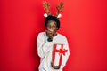 Young african american man wearing deer christmas hat holding gift laughing and embarrassed giggle covering mouth with hands,