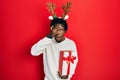 Young african american man wearing deer christmas hat holding gift peeking in shock covering face and eyes with hand, looking