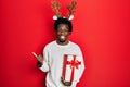 Young african american man wearing deer christmas hat holding gift pointing to the back behind with hand and thumbs up, smiling Royalty Free Stock Photo