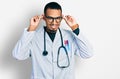 Young african american man wearing doctor uniform and stethoscope smiling pulling ears with fingers, funny gesture