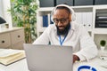 Young african american man wearing doctor uniform using laptop and headphones working at clinic Royalty Free Stock Photo