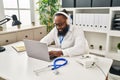 Young african american man wearing doctor uniform using laptop and headphones working at clinic Royalty Free Stock Photo