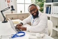 Young african american man wearing doctor uniform using laptop and headphones working at clinic Royalty Free Stock Photo