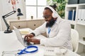 Young african american man wearing doctor uniform using laptop and headphones working at clinic Royalty Free Stock Photo